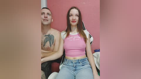 Video of a shirtless, tattooed man with a muscular build and a woman in a pink crop top and blue jeans, sitting side by side on a couch against a pink wall.