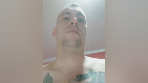 A video of a bald, light-skinned man with a trimmed beard and tattoos on his chest and shoulder, wearing a shirt, standing against a red wall in a room with white trim.