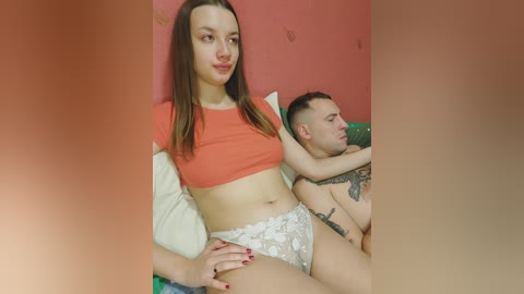 A video of a young Caucasian woman with long brown hair, wearing an orange crop top and white lace panties, sitting next to a tattooed man in bed.