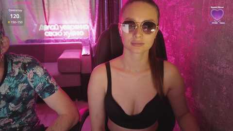 Video of a woman with long hair, wearing black lingerie, glasses, and a pink backdrop. She sits in a dimly lit room with neon signs and a man in a tropical shirt.
