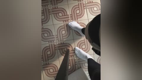 Media: Video of a person's feet with white socks, standing on a tiled floor with a geometric brown pattern. The background is blurred, and the perspective is from above.