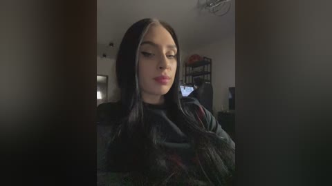 Media: Video of a young woman with pale skin, long black hair, and a dark black cloak, seated indoors with a dimly lit room and visible furniture in the background.