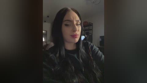 Video of a woman with long, straight black hair, wearing a black jacket, standing in a dimly lit room with shelves and a lamp in the background.