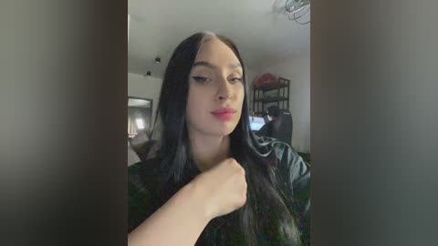 Video of a young woman with long black hair, fair skin, and full lips, wearing a black jacket, standing in a dimly lit room with a TV and bookshelf in the background.