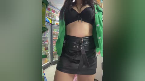 Media: Video of a woman with long black hair in a black bra and green jacket, wearing a black high-waisted leather skirt with a belt, standing in front of a brightly lit convenience store refrigerator.