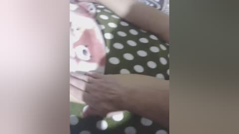 A video of a person's hands holding a smartphone, with a green polka-dotted shirt and a pink shirt with a cartoon character. The background is blurred, with indistinct colors.