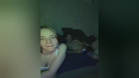 A dimly lit video shows a blonde woman in a white tank top lying on a dark couch, smiling towards the camera. A man, shirtless, lies beside her, also smiling.