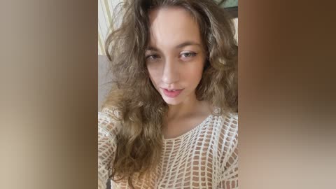 Video of a fair-skinned woman with long, wavy brown hair, wearing a cream-colored, crocheted top. She has a neutral expression and is indoors, with blurred, warm-toned background.