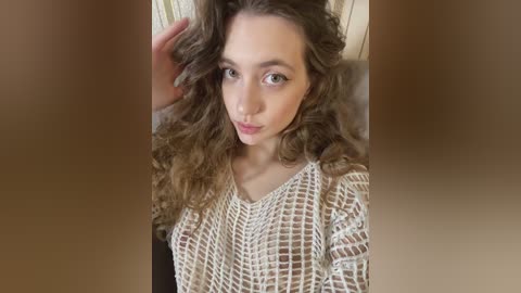 Video of a young woman with fair skin and wavy, light brown hair, wearing a beige, knitted sweater, posing indoors against a blurred background.