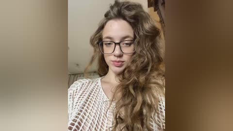 A video of a young woman with long, wavy brown hair, wearing black-rimmed glasses and a white crochet top. She stands in an indoor setting with beige walls and a wooden staircase in the background.