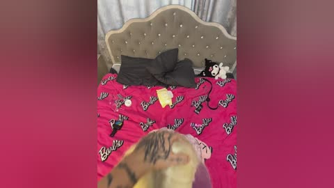 Media: Video of a messy bedroom with a pink bedspread featuring \"Barbie\" text, a beige tufted headboard, a gray pillow, a stuffed cat, and a yellow envelope.