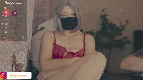 A video of a young Asian woman with light skin and shoulder-length gray hair, wearing a black mask, red lace bra, and shorts, seated on a chair in a dimly-lit, cozy room.