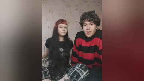 Media: Video of a young, pale-skinned woman with long, straight red hair and a black t-shirt, seated beside a curly-haired, dark-skinned man in a red and black striped sweater, both in a dimly lit room with a patterned wallpaper.