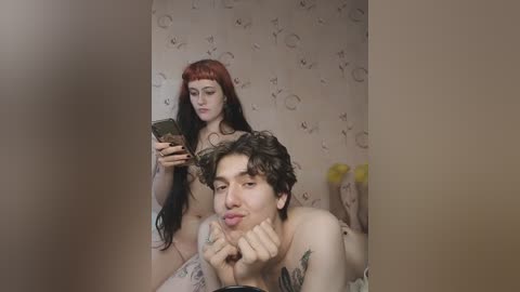 Media: Video of a naked, tattooed young man with long, curly brown hair, resting his chin on his hand, and a naked woman with long, straight red hair, standing behind him. Both are in a beige-patterned room.