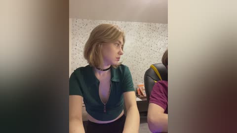 Video of a young woman with shoulder-length blonde hair, wearing a teal, low-cut, short-sleeved top, and a black choker, seated in a modern, dimly-lit room with speckled white walls and a gaming chair in the background.