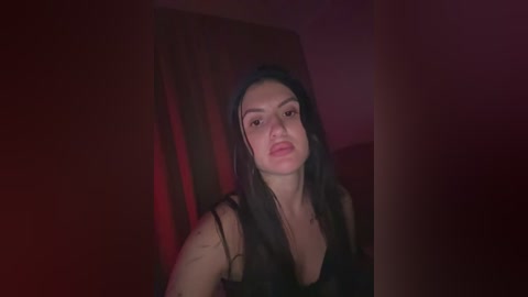 Video of a young woman with long black hair, light skin, and dark eyes, wearing a black top, standing in a dimly lit room with red walls.