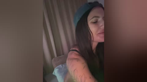 A video of a young woman with long, dark hair, wearing a blue hat and a black top, leaning against a beige curtain, partially obscured by another person's arm.