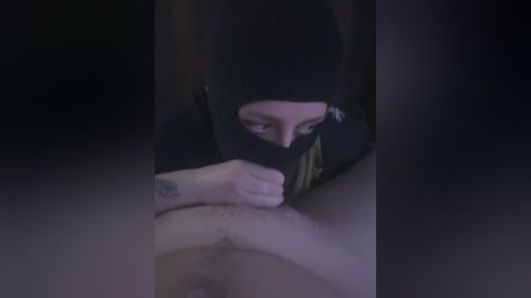 A dimly lit video shows a woman with dark hair and a black beanie, wearing a black shirt, kneeling to perform oral sex on a man's erect penis.
