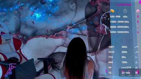 A video of a woman with long brown hair, wearing a red top, seated in front of a large, detailed, digitally rendered painting on a computer screen.