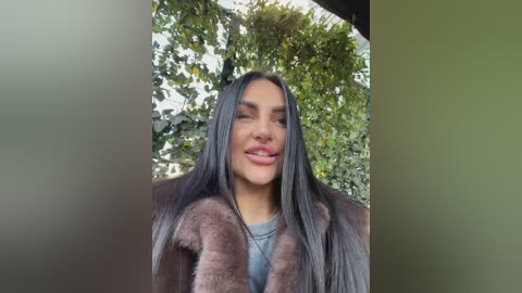 Media: Video of a smiling woman with long, straight black hair, wearing a fur-trimmed coat, standing in front of a lush green ivy-covered wall.