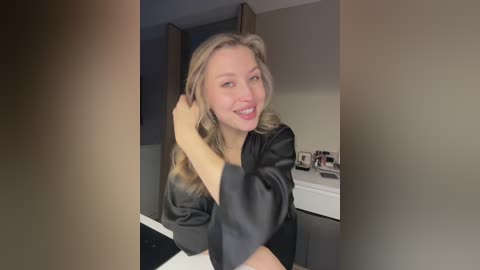 Media: Video of a smiling Caucasian woman with wavy blonde hair, wearing a black satin robe, seated in a modern bathroom with white countertops and wooden cabinets.