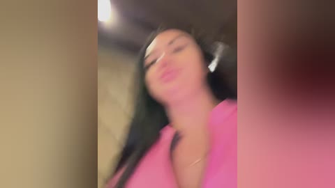 A blurry video of a young woman with long black hair, wearing a pink shirt, taken in a dimly lit room.