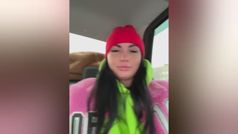 A video of a young woman with long black hair and fair skin, wearing a bright red beanie and a neon green jacket, sitting in a car.