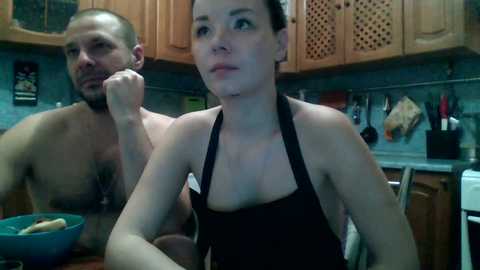 A video shows a shirtless, muscular man with a shaved head and a tattooed chest, standing behind a woman in a black apron, in a kitchen with wooden cabinets, utensils, and a blue bowl.