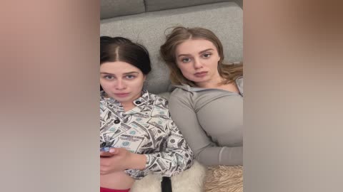 Media: Video of two young women with fair skin, dark hair, and light gray sweaters, lying on a gray sofa; one holds a controller, the other rests her head on her arm.