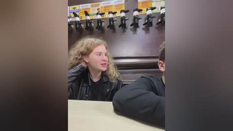 Media: Video of a young woman with curly blonde hair, wearing a black leather jacket, sitting in a TV studio with a backdrop of yellow and white advertisements and multiple cameras.