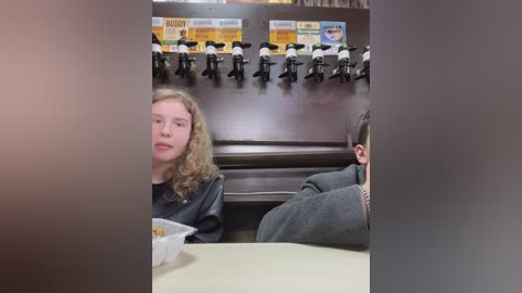 Media: Video of a young girl with curly blonde hair in a black leather jacket, eating from a white container at a fast-food restaurant. Behind her, a soda dispenser with toy figurines is mounted on the wall.