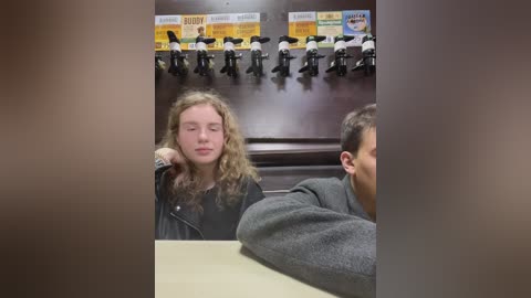 Media: A video showing a young girl with curly blonde hair, wearing a black jacket, seated at a table in a classroom with a row of black chairs and a poster behind her.