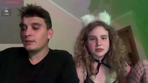 Media: Video of a man with short dark hair in a black shirt, and a woman with curly brown hair, white bunny ears, and fishnet stockings, indoors.