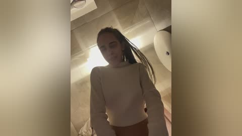 Media: Video of a young woman with light skin, wearing a beige sweater and high-waisted brown pants, standing in a beige-tiled bathroom with a round mirror and a towel rack.