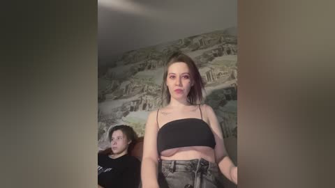 A video featuring a young woman with fair skin and short brown hair, wearing a black crop top and high-waisted jeans, standing in front of a wall mural depicting a historical cityscape. A young boy with curly hair sits behind her.