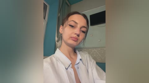 A video of a young woman with light skin, brown hair pulled back, wearing a white button-up shirt and black eyeliner, standing in a teal-painted room with a white wall and a mirror.