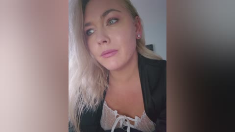 Video of a blonde woman with fair skin, wearing a black top with a lace trim, looking contemplative, framed by blurred figures.