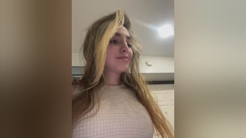 Media: Video of a young woman with long, messy blonde hair, wearing a light pink ribbed sweater, standing indoors with white cabinets and a ceiling light in the background.