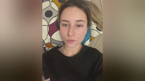 A video of a young woman with light skin and brown hair, wearing a black T-shirt, standing against a background with a colorful, abstract pattern.