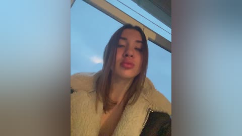 Media: A video of a young woman with light skin and straight, shoulder-length brown hair, wearing a beige, fluffy jacket, taken indoors near a large window with a cloudy sky visible in the background.