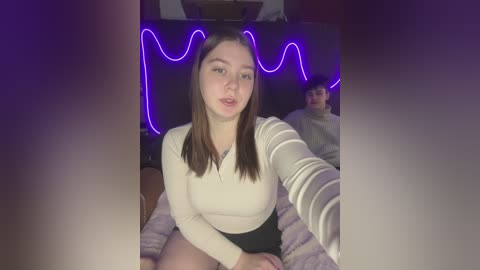 Media: Video of a young Caucasian woman with long brown hair, fair skin, and a slender build, wearing a cream long-sleeved top and black skirt, sitting on a bed with a purple blanket. A young man sits in the background.