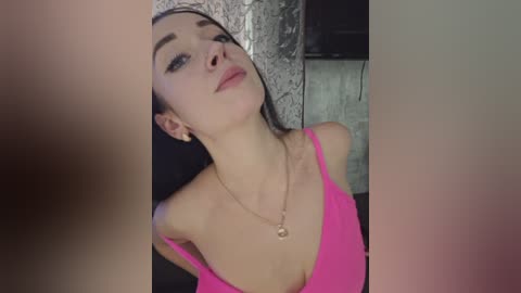 Video of a young woman with fair skin, long dark hair, wearing a bright pink off-shoulder top, and a delicate necklace. Background features a textured, metallic wall.