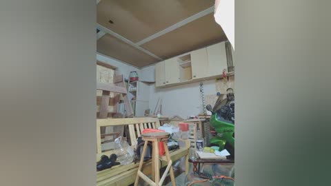 Video of a cluttered, unfinished home workshop with exposed walls, unfinished wood cabinets, a green tarp, tools, and assorted materials scattered on wooden shelves and workbenches.