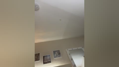 Media: A video of a modern, minimalist room with a white ceiling, a circular light fixture, and three framed abstract art pieces. The walls are beige, and a white-framed mirror is partially visible on the right.