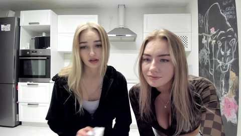 Media: Video of two young blonde women in a modern kitchen, one with a white tank top and black jacket, the other with a brown jacket, both looking towards the camera.