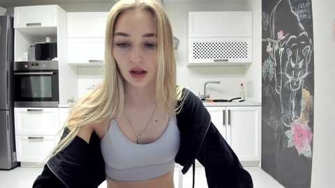 Media: Video of a young, fair-skinned woman with long blonde hair, wearing a gray sports bra and black jacket, in a modern kitchen with white cabinets, black oven, and chalkboard wall.