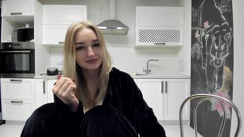 Media: Video of a blonde woman in a black robe, sitting in a modern white kitchen with stainless steel appliances and a floral mural on the wall.