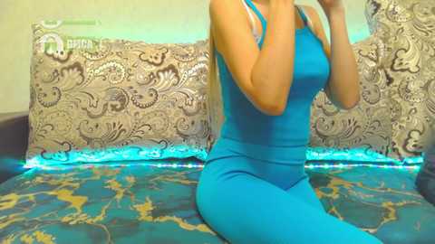 Media: Video of a fair-skinned woman with long blonde hair, wearing a tight, bright blue sleeveless dress, sitting on a bed with a blue and gold patterned duvet and ornate pillowcase in a light-colored room.
