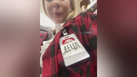 Video of a blonde woman with fair skin wearing a red and black plaid jacket, a white shirt, and an ID badge with \"AEA\" and a red logo. The background is blurry.