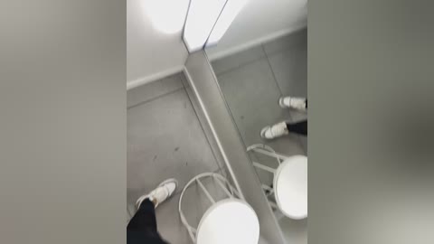 Media: A video of a narrow restroom stall with a bright overhead light, revealing a white toilet, a white sink, and a white toilet paper dispenser. The floor is gray, and the mirror reflects the scene, showing a person in white shoes.